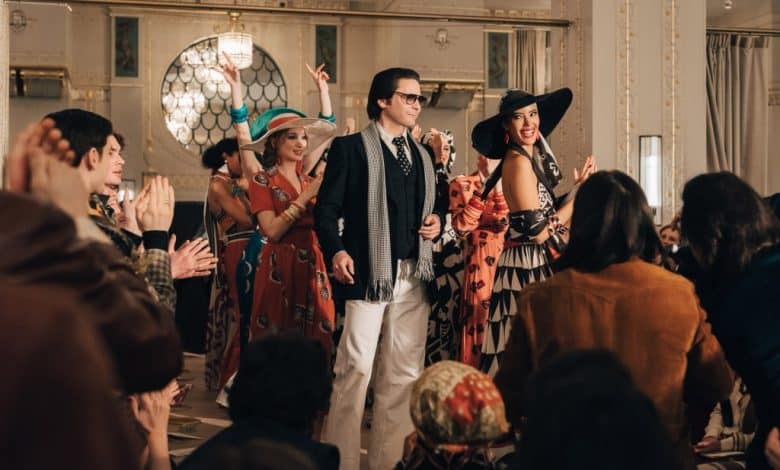 A joyful group dressed in vintage fashion, inspired by the Karl Lagerfeld Series: Beyond Biopic, including a man in sunglasses and a woman in a wide-brim hat, walk through a crowd cl