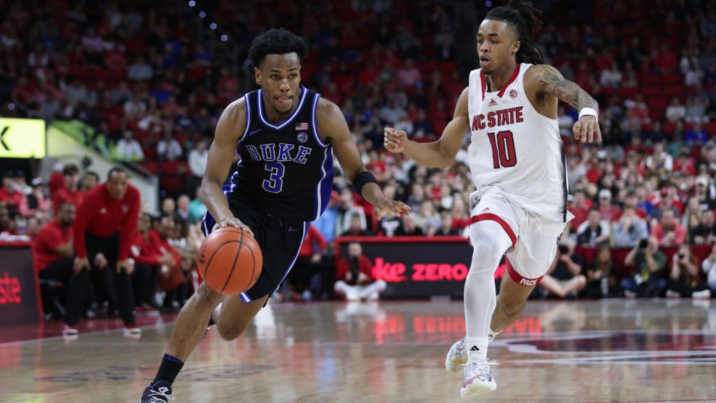 Two college basketball players in action during the 2024 NCAA Elite Eight game. A Duke player in a blue uniform, number 3, dribbles the ball quickly past an N.C. State player