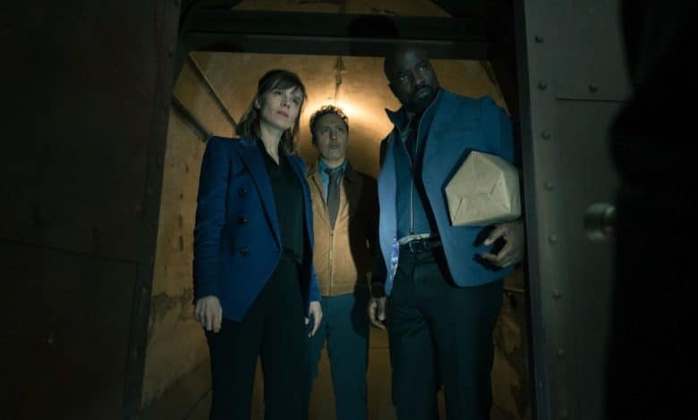 Three people stand inside a dim, industrial-looking space, looking wary—a woman in a blue jacket, a man in a suit holding a box, and a tall man in a blue shirt. Their expressions