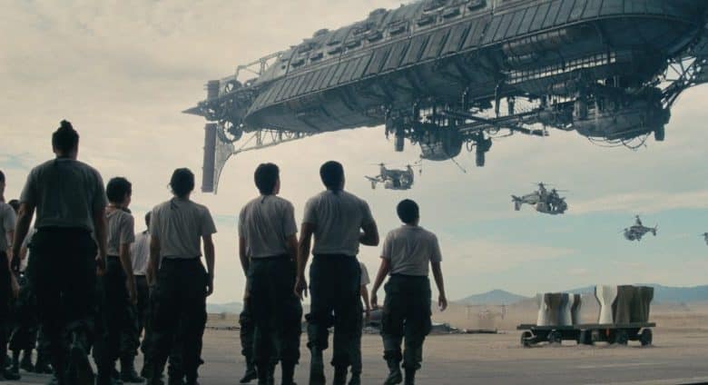 A group of people in military uniforms watch as a large, futuristic hovercraft and several smaller flying crafts maneuver above the vast desert landscape of California.