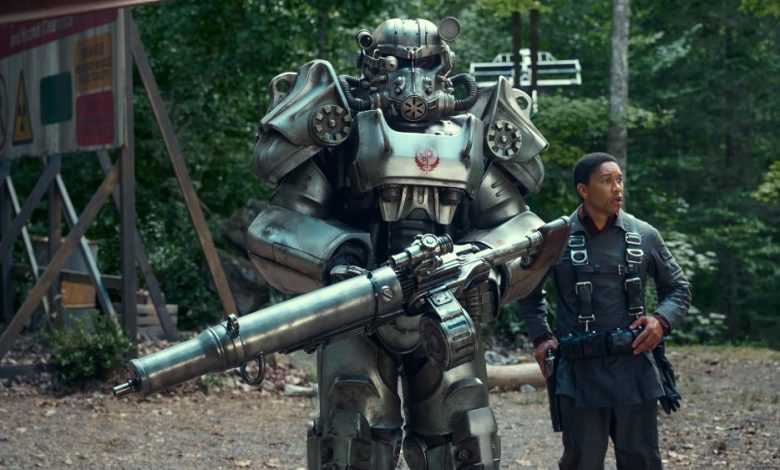 A man in tactical gear stands next to a large, metallic robot equipped with armor and a big gun, in a forest near a signboard announcing the 'Fallout' series' new release date on