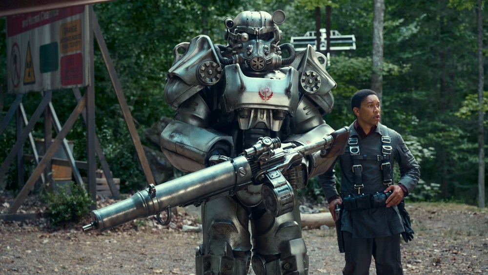 A man in tactical gear stands next to a large, metallic robot equipped with armor and a big gun, in a forest near a signboard announcing the 'Fallout' series' new release date on