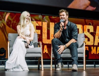 A man and a woman sit on stage at a promotional event for "Furiosa: A Mad Max Saga Unveiled." The woman in a white dress smiles while seated, and the man,