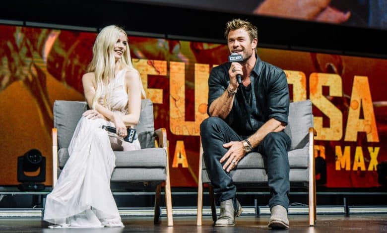 A man and a woman sit on stage at a promotional event for "Furiosa: A Mad Max Saga Unveiled." The woman in a white dress smiles while seated, and the man,