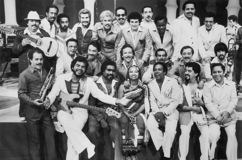 A black and white photo of a large group of smiling musicians from Fania Records' 60th Anniversary, including singers and various instrumentalists with guitars, saxophones, and trumpets. They