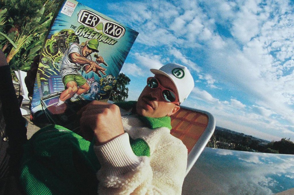 A person wearing a green and white cap, sunglasses, and a matching jacket poses with a comic book titled "Ferxxo the Green Man," held prominently in the foreground against a scenic, sun