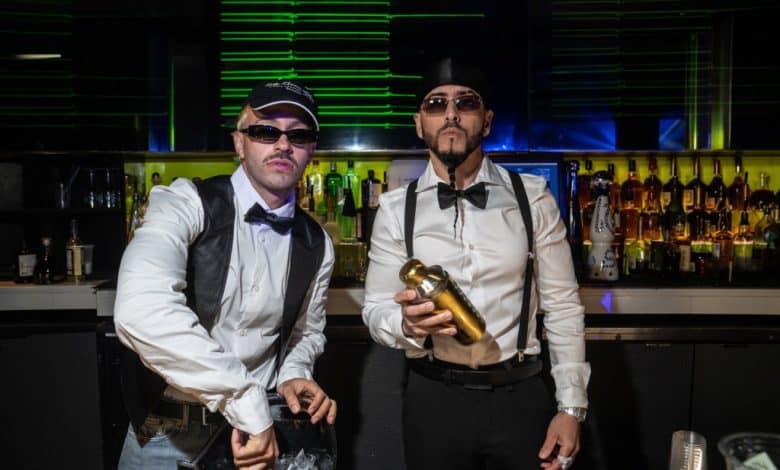 Two stylish bartenders posing behind a bar, one shaking a cocktail shaker. Both wear suspenders, bow ties, and caps, with a backdrop of liquor bottles and glowing green lights.