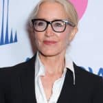 A woman with blonde hair and glasses, wearing a black blazer over a white ruffled blouse, smiles at the Season 2 premiere of 'Criminal Minds: Evolution' with a step and repeat