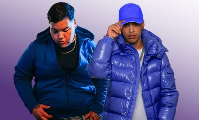 Two men posing confidently against a purple background. The man on the left wears a blue hoodie, and the one on the right wears a blue puffer jacket and blue hat, adjusting the brim.