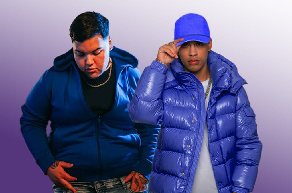 Two men posing confidently against a purple background. The man on the left wears a blue hoodie, and the one on the right wears a blue puffer jacket and blue hat, adjusting the brim.