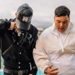 Two young men standing by a pool with a scenic backdrop of a cloudy sky and green fields. One wears a black jacket and cap, covering his face, the other in a white jacket looks down at
