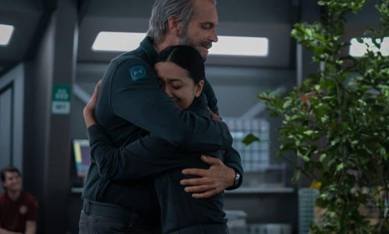 Two people, an older man and a younger woman, embrace warmly in a dimly lit room with futuristic decor. The man is smiling gently as he hugs the woman, who appears comforted by the
