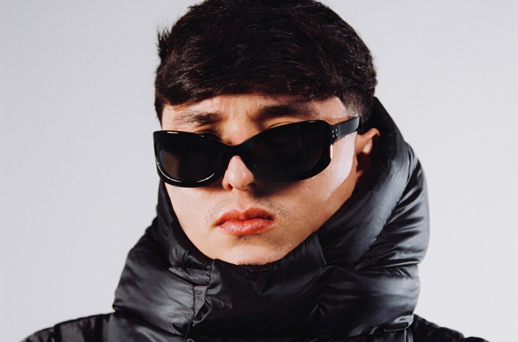 A young man wearing oversized black sunglasses and a glossy black puffer jacket looks directly at the camera with a neutral expression, against a plain light background, in the promotional materials for the 2024 U