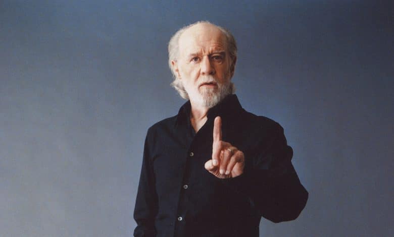 An elderly man with white hair and a beard, dressed in a black shirt, sternly holds up one finger in a gesture of emphasis or warning, against a soft blue background, symbolizing the recent