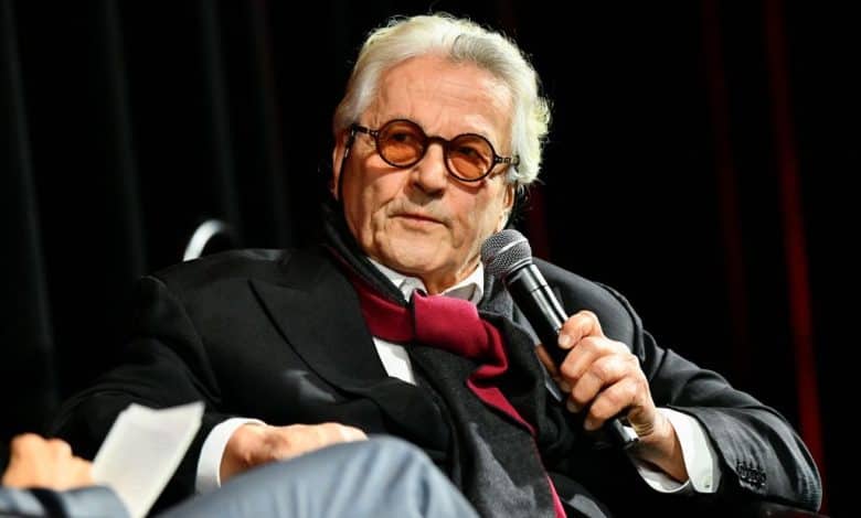 A distinguished older man with white hair and a mustache speaks into a microphone while seated on stage. He wears a black suit, white shirt, red bow tie, and round glasses, with a thoughtful