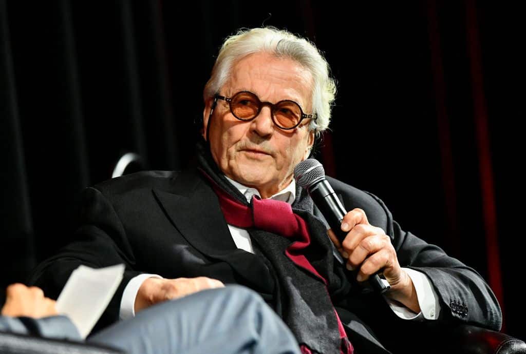 A distinguished older man with white hair and a mustache speaks into a microphone while seated on stage. He wears a black suit, white shirt, red bow tie, and round glasses, with a thoughtful