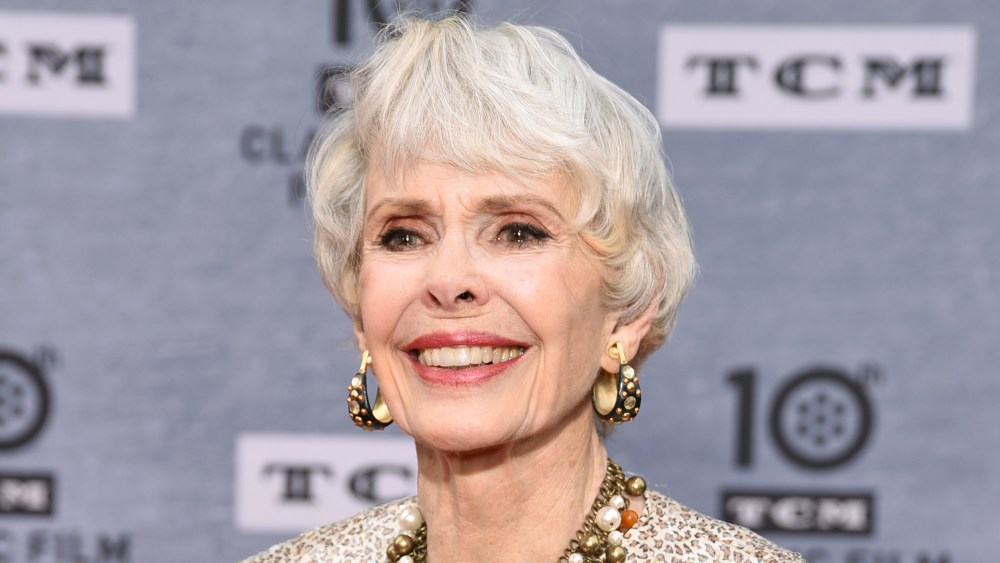 Elderly woman with short white hair and bright smile, wearing large gold earrings and a beige embellished dress, posing in front of a grey, logo-patterned backdrop.