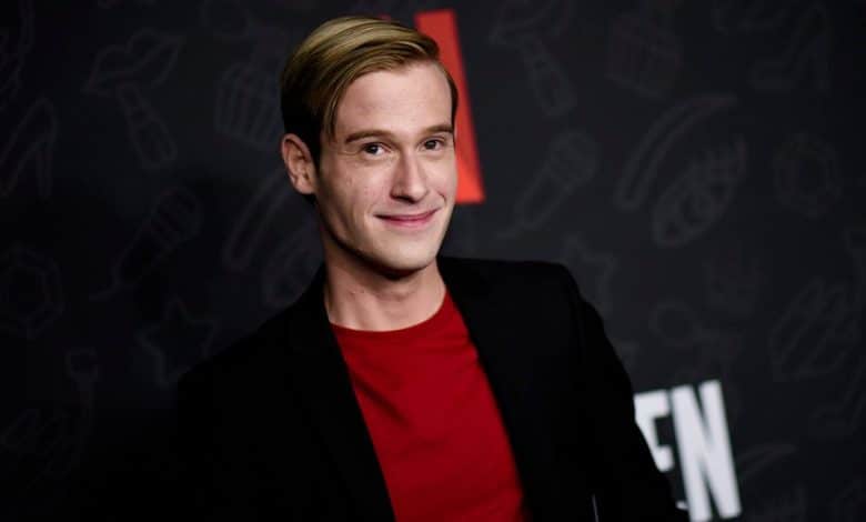A man with short blond hair, smiling at the camera, wearing a red shirt under a black blazer at a media event with a background featuring doodle art and the letter "n," titled Tyler