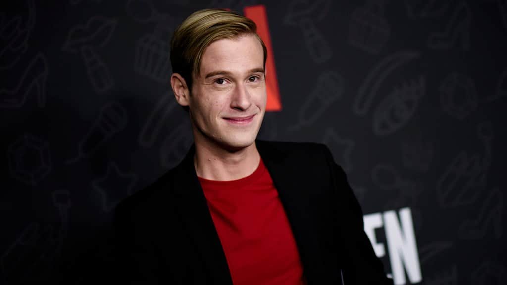 A man with short blond hair, smiling at the camera, wearing a red shirt under a black blazer at a media event with a background featuring doodle art and the letter "n," titled Tyler