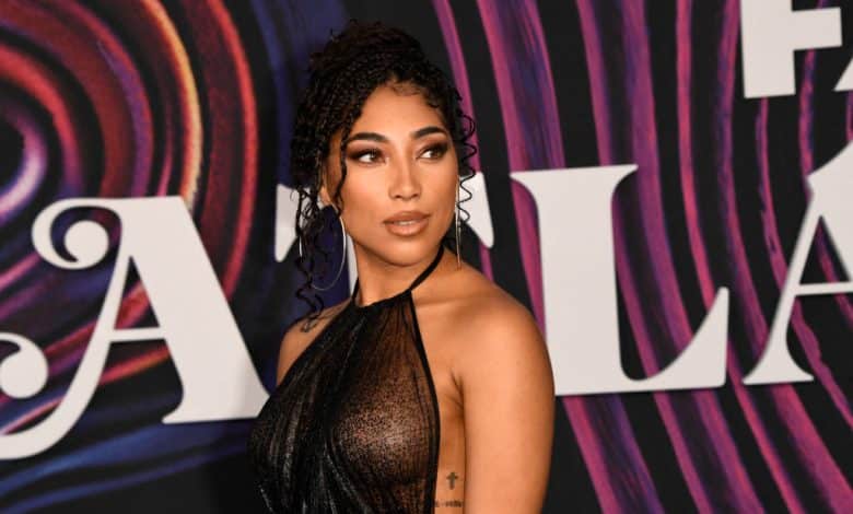 A woman with an elegant updo and side-swept braids poses at a gala event. She wears a shimmering, one-shoulder black gown against a colorful, swirl-patterned backdrop