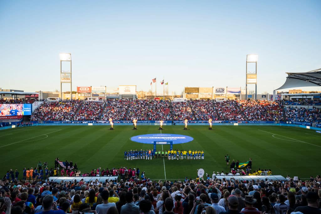 A vibrant soccer match featuring the USWNT in the 2024 SheBelieves Cup begins at sunset in a stadium filled with spectators. Two teams line up in the center, beneath towering floodlights