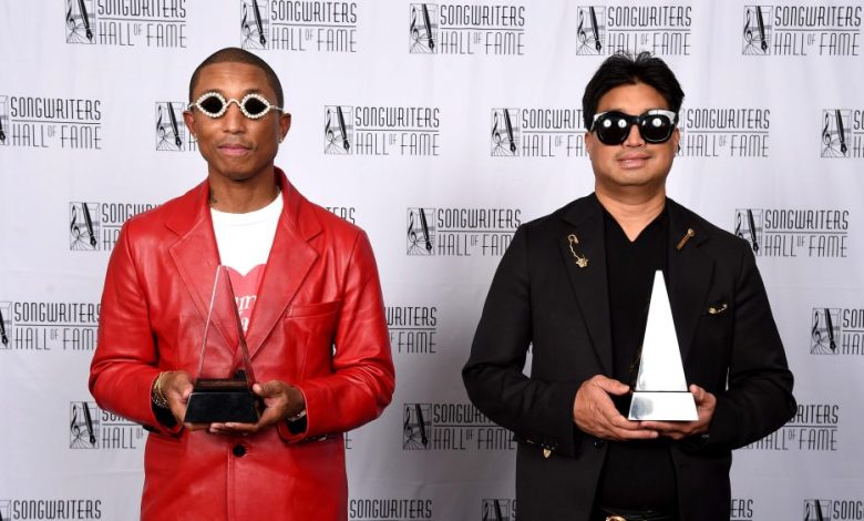 Two men stand against a logo backdrop, each holding an award. The left man, Williams, wears a red jacket and sunglasses; the right man sports a black jacket and sunglasses. Both are smiling.
