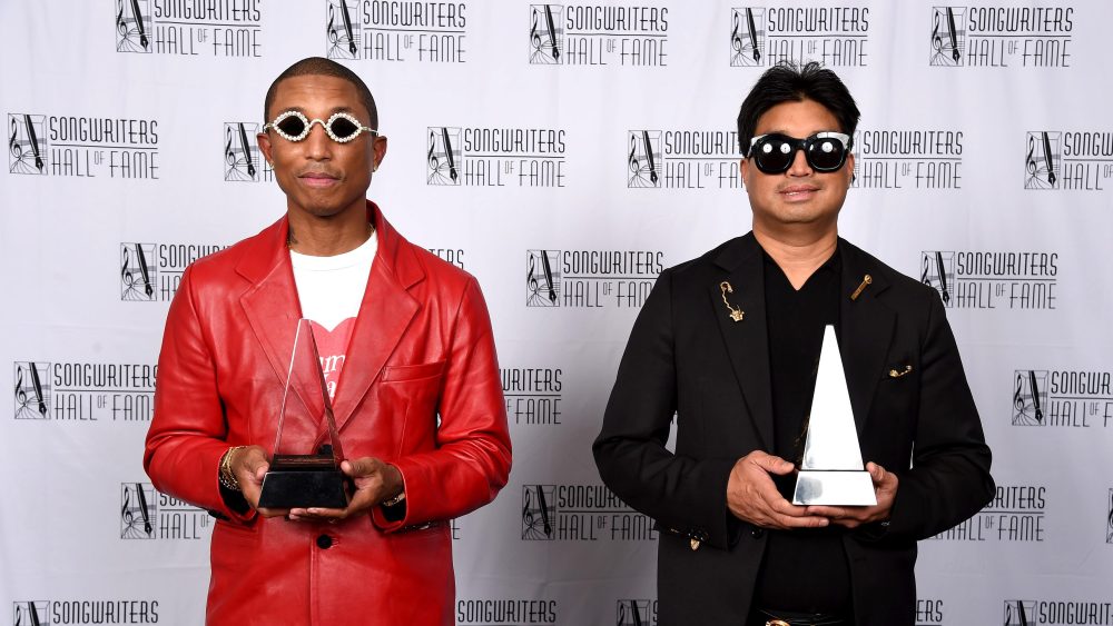 Two men stand against a logo backdrop, each holding an award. The left man, Williams, wears a red jacket and sunglasses; the right man sports a black jacket and sunglasses. Both are smiling.