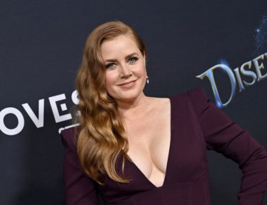Amy Adams poses at a Disney event, wearing a deep purple dress with a plunging neckline. Her hair is styled in waves, and she smiles confidently at the camera.