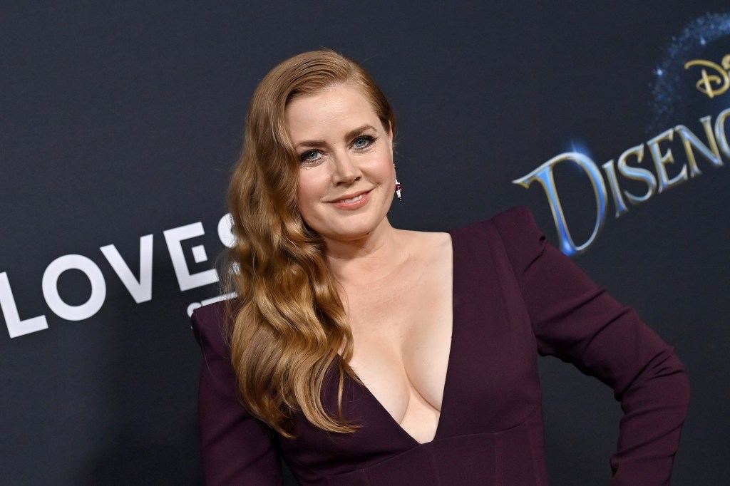 Amy Adams poses at a Disney event, wearing a deep purple dress with a plunging neckline. Her hair is styled in waves, and she smiles confidently at the camera.