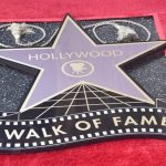 A Hollywood Walk of Fame star on a red carpet, featuring the iconic emblem with a movie camera in the center. The star is partially covered by two small circular furry objects at the top. The