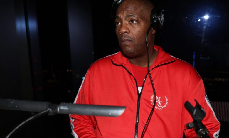 A man in a red jacket with a DJ Mister Cee logo, wearing headphones, stands by a microphone in a dimly lit area, looking to the side with a focused expression.