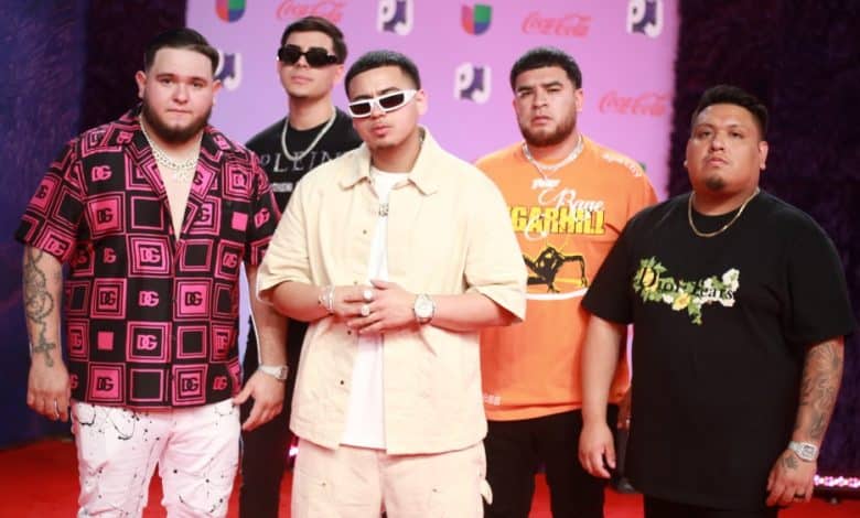 Five men pose on a red carpet at a Fuerza Regida event. They are dressed in casual, stylish attire, ranging from graphic tees to patterned shirts, and accessories like sunglasses and
