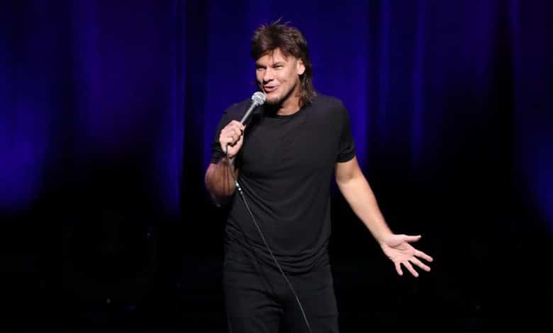 A male comedian performing on stage, holding a microphone, wearing a black t-shirt and jeans. He gestures animatedly with his right hand as he speaks, with a focused expression under a spotlight against