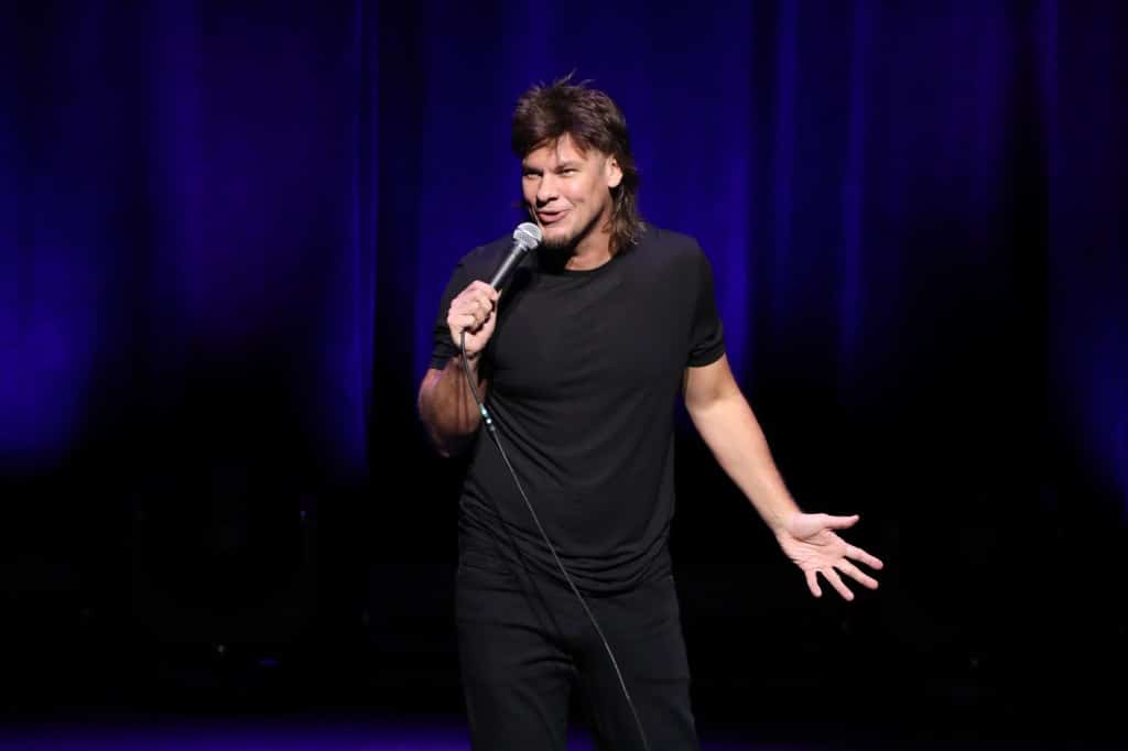 A male comedian performing on stage, holding a microphone, wearing a black t-shirt and jeans. He gestures animatedly with his right hand as he speaks, with a focused expression under a spotlight against