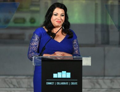 A woman stands at a podium giving a speech at an evening event. She wears a blue lace dress and smiles confidently. The podium has the Hispanic Media Coalition's Campaign for Positive Change in Industry logo on