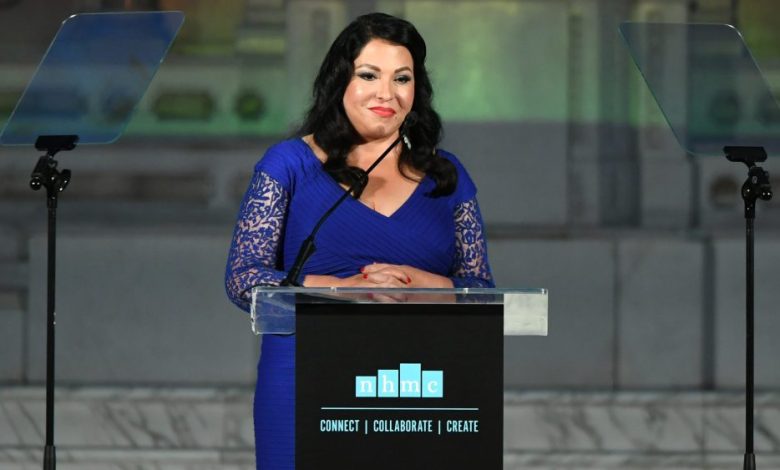 A woman stands at a podium giving a speech at an evening event. She wears a blue lace dress and smiles confidently. The podium has the Hispanic Media Coalition's Campaign for Positive Change in Industry logo on