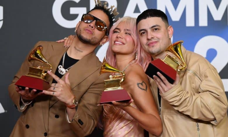 Three musicians pose with Latin GRAMMY awards, smiling at the camera. They have unique hairstyles and vibrant attire, celebrating their success at the event. The backdrop features the Grammy logo.