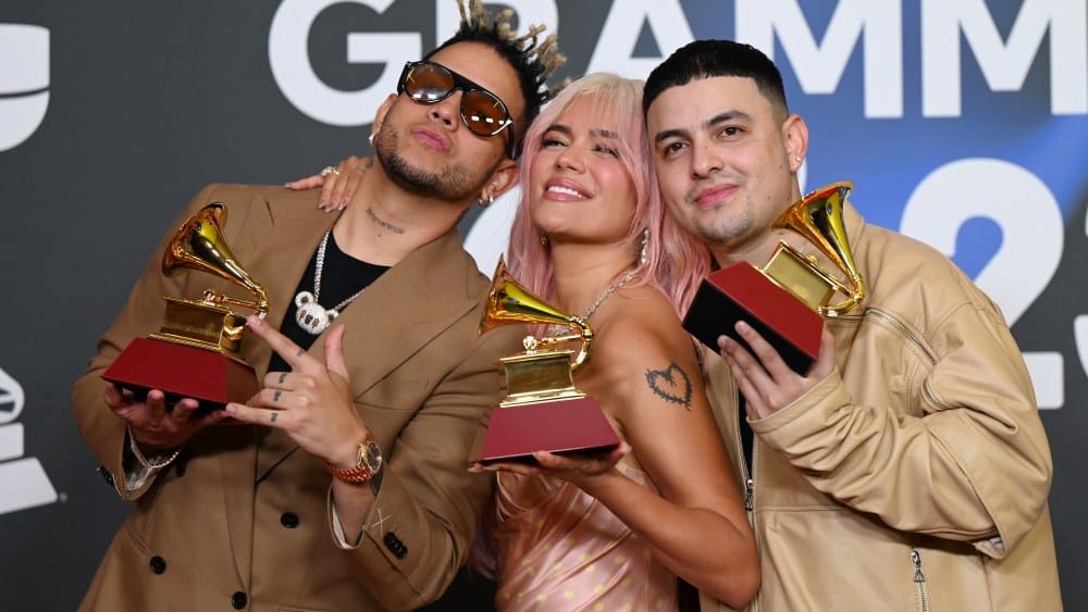Three musicians pose with Latin GRAMMY awards, smiling at the camera. They have unique hairstyles and vibrant attire, celebrating their success at the event. The backdrop features the Grammy logo.