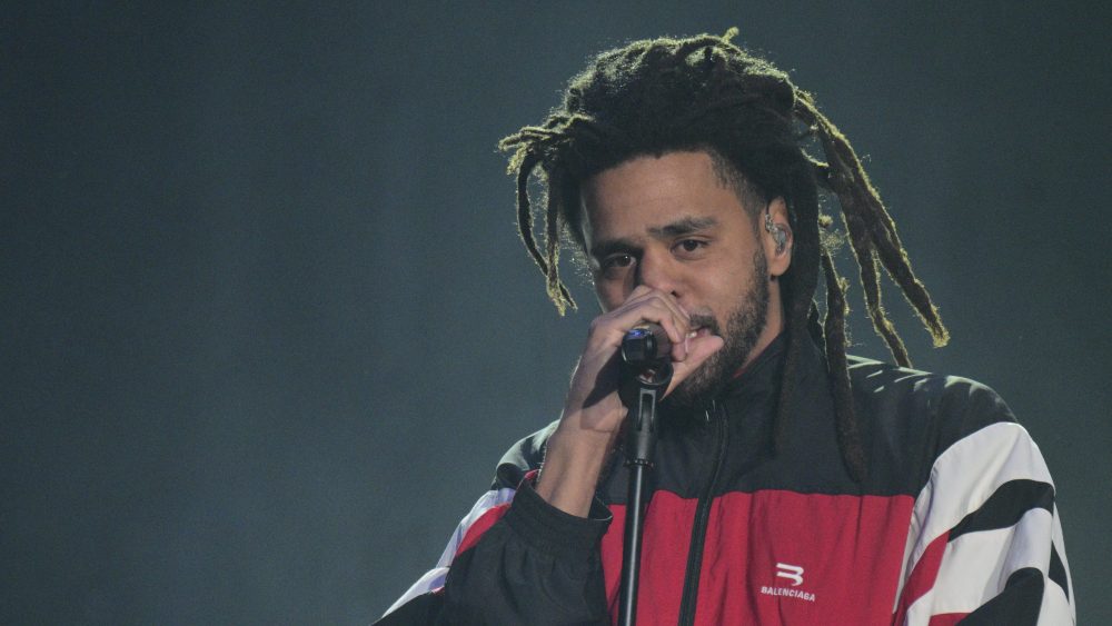 A male singer with dreadlocks, wearing a red, white, and black striped jacket, sings into a microphone on a dimly lit stage, showing intense expression and feeling horrible regrets.
