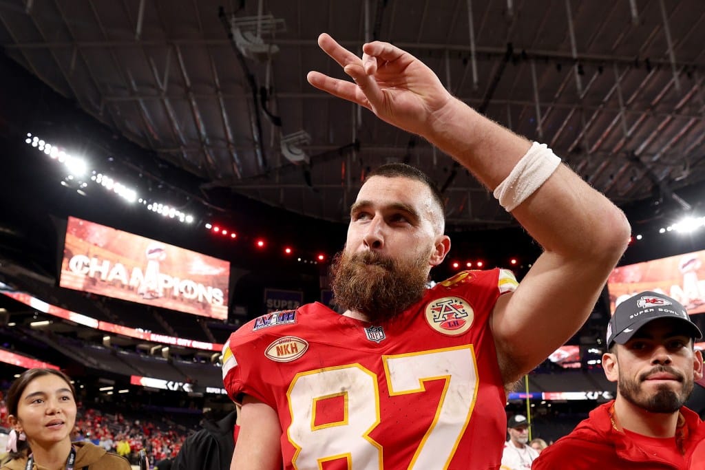 A bearded football player, Travis Kelce, in a red Kansas City Chiefs jersey with number 87 celebrates by pointing upwards, in a crowded stadium with a "champions" sign in the background