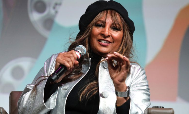 A woman wearing a silver blazer and black beret speaks into a microphone, gesturing with her hands. She sits onstage with a blurred colorful background, promoting the 'Foxy: My Life in