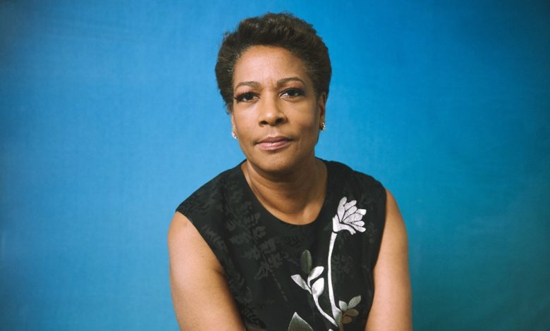 A poised middle-aged black woman, possibly reminiscent of director Dawn Porter, with short hair looks directly at the camera, wearing a black sleeveless top with a white floral design, against a blue backdrop.