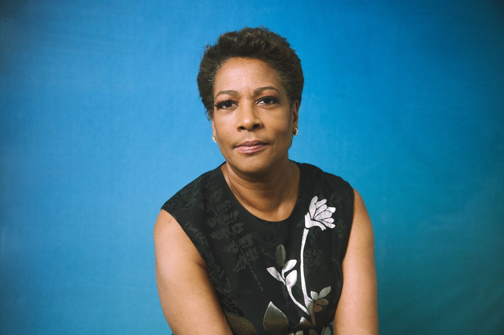 A poised middle-aged black woman, possibly reminiscent of director Dawn Porter, with short hair looks directly at the camera, wearing a black sleeveless top with a white floral design, against a blue backdrop.