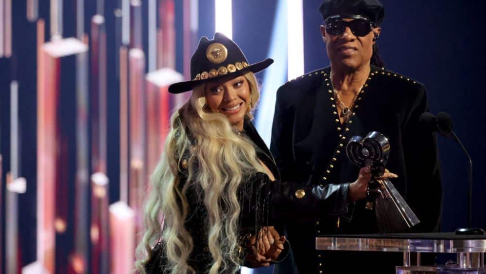 Two celebrities on a stage at an awards ceremony, smiling and holding a trophy. One wears a decorative hat and black outfit, and the other sports a cowboy hat with a chain necklace.