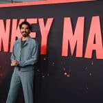 A man in a gray suit stands confidently in front of a large red banner with the words "Monkey Man" in bold white letters. His hands are clasped in front, and he smiles slightly at