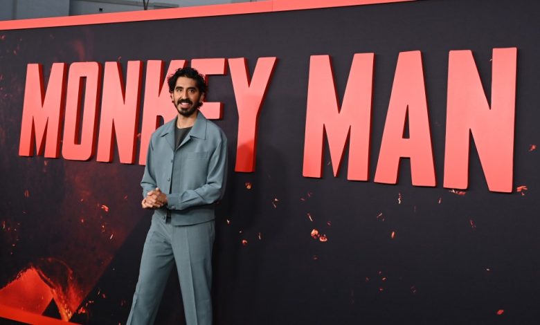 A man in a gray suit stands confidently in front of a large red banner with the words "Monkey Man" in bold white letters. His hands are clasped in front, and he smiles slightly at