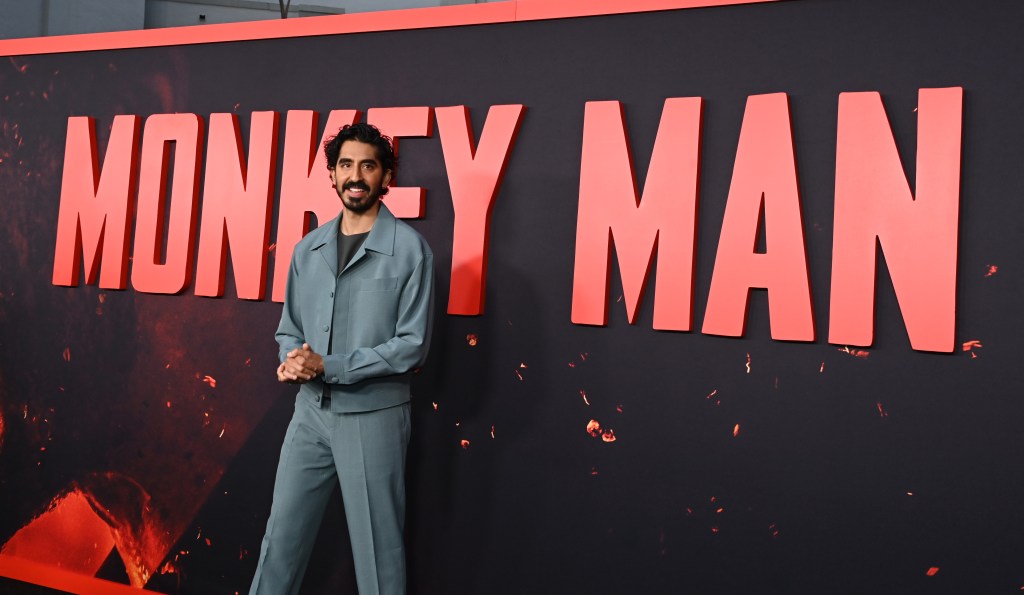 A man in a gray suit stands confidently in front of a large red banner with the words "Monkey Man" in bold white letters. His hands are clasped in front, and he smiles slightly at