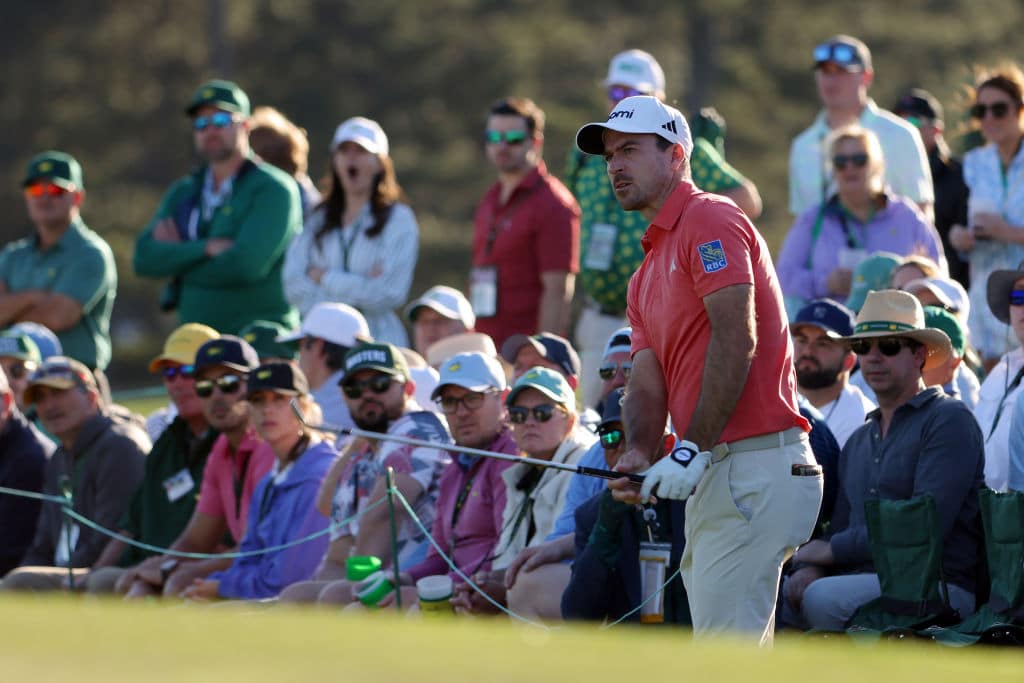 A golf player in a red shirt and white cap watches his shot intently amid a gallery of spectators in various shades of attire, mostly greens and neutrals, during ESPN's Masters Tourney Rounds