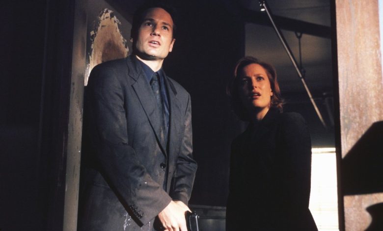 Two people, a man in a dark suit and a woman in a black coat, standing in an 'X-Files'-like dimly lit room with unease, looking off to the side as