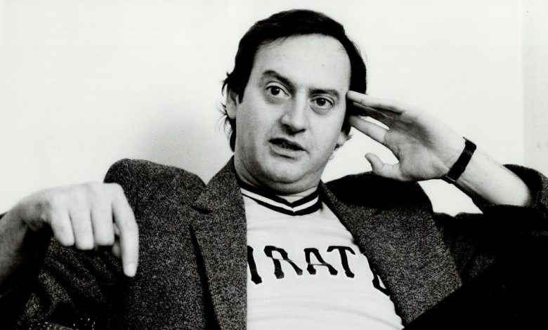A black-and-white photo of a middle-aged man with a thoughtful expression, touching his forehead, sitting with one arm draped over a chair. He wears a sports t-shirt under a blazer as stars